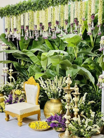 A close-up of a classic Seemantham setup, highlighting the rich texture of the banana leaf backdrop. The scene is completed with an ornate chair, brass lamps, and vases of fresh tuberose.