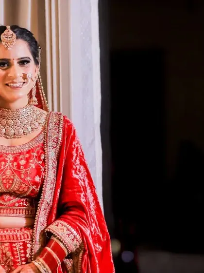The bride looks radiant in her red wedding lehenga. This colour portrait captures her beauty and happiness against a dark, contrasting background.