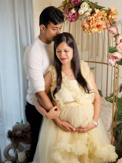 This candid moment shows the couple sharing a quiet connection. He gently kisses her forehead as they both cradle the bump, set against the warm, floral-adorned boho backdrop.