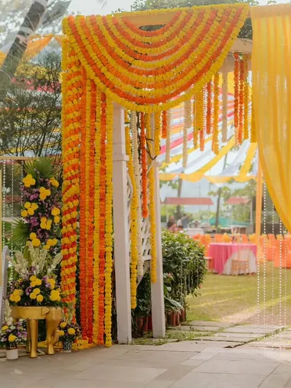 The entrance to a Haldi celebration, featuring a white picket fence gate adorned with marigold garlands and lush floral arrangements. A welcome sign greets the guests, setting a warm and inviting tone.