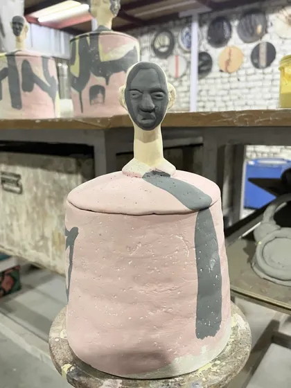 A finished 'Human Jar' before its final firing. The piece is coated in colored slips, with a sculpted head as the handle, blending functional pottery with unique sculptural elements.