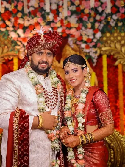 A classic stage portrait of a smiling couple, set against a backdrop of rich, golden, and red floral decorations typical of a grand Indian wedding.