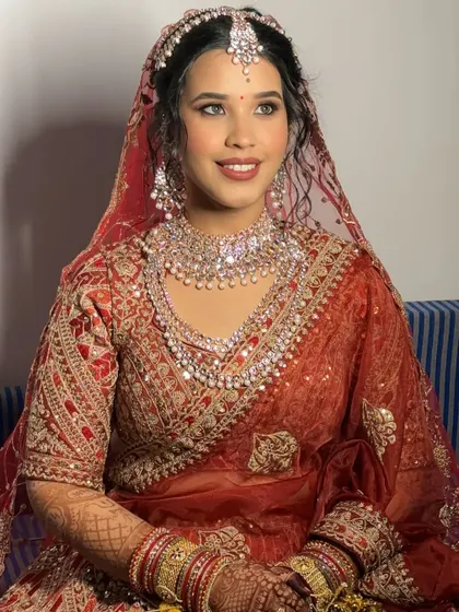 A beautiful seated portrait of the bride in her red lehenga. The makeup features classic elements like defined eyes and a bold lip, perfect for a traditional ceremony.