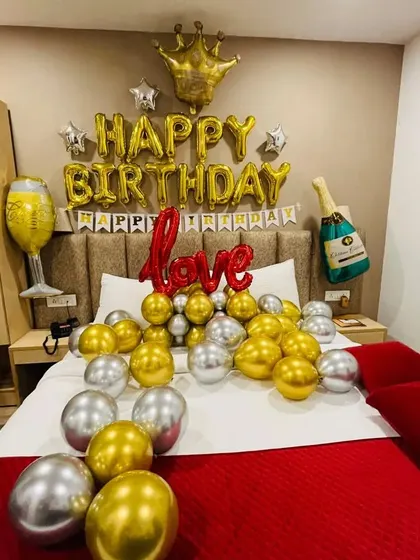 A celebratory birthday bedroom decoration. The bed is filled with gold and silver balloons, and the wall features 'Happy Birthday' foil letters, a crown, and champagne-themed balloons for a fun, festive feel.