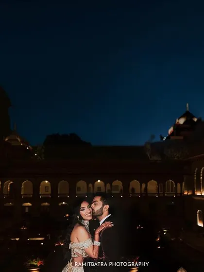 A romantic nighttime portrait of the couple at Rambagh Palace, with the groom kissing the bride's cheek.