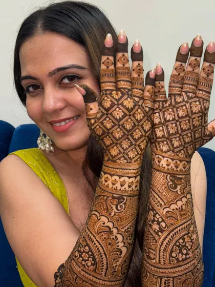 A happy bride showcasing her intricate grid-patterned mehendi that extends up her arms, perfect for her wedding day.