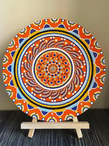 A smaller dot mandala from my collection, displayed on an easel. This piece uses a bright orange base with blue and yellow accents, creating a warm and energetic design.