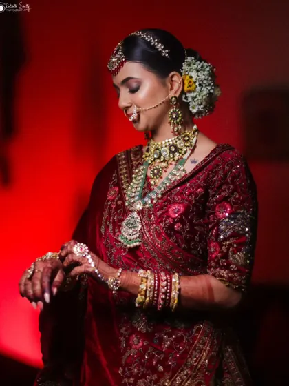 A side profile of the bride in her red lehenga, checking her watch. This candid shot captures a moment of anticipation, with the floral hairpiece adding a soft, traditional touch.
