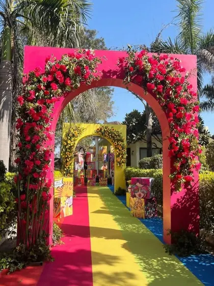 A vibrant and colorful entrance arch for a daytime wedding event. The bright pink and yellow structure is adorned with red roses, setting a festive mood.