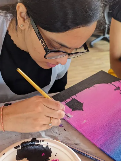 A participant carefully painting a nightscape on canvas, adding silhouettes of trees against a colorful sky.