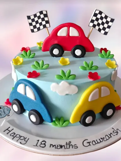 A simple and sweet car-themed cake for an 18-month celebration. It features three colorful fondant cars on a light blue whipped cream base.