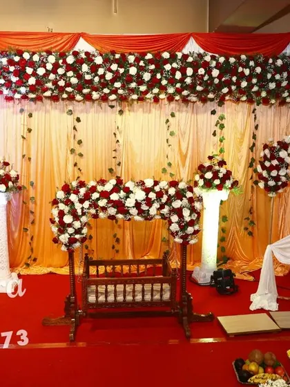 A beautiful naming ceremony decoration in classic red and white. The setup includes a traditional wooden cradle decorated with a floral arch, and a personalized photo frame of the baby.