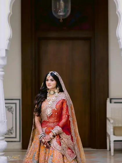 A regal portrait of a bride in her orange and red lehenga, framed by the ornate white arches of a palace in Udaipur.