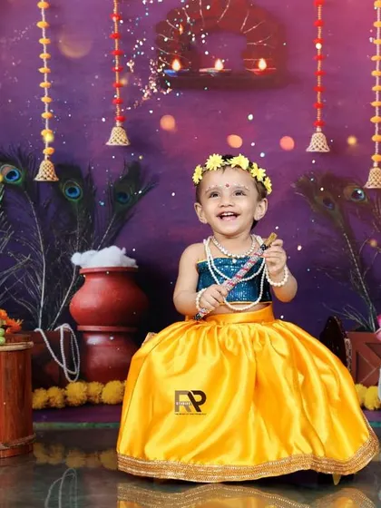A smiling baby girl dressed in a beautiful yellow and blue outfit for a Radha-themed photoshoot. The festive backdrop with marigolds and peacock feathers is perfect for celebrating Janmashtami.