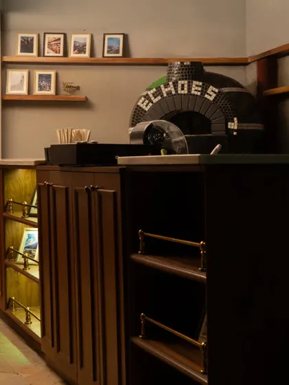Behind the bar, a mosaic-tiled pizza oven proudly displays the Echoes name. The design combines functionality with character, featuring warm wooden cabinetry and brass details for a sophisticated yet cozy ambiance.