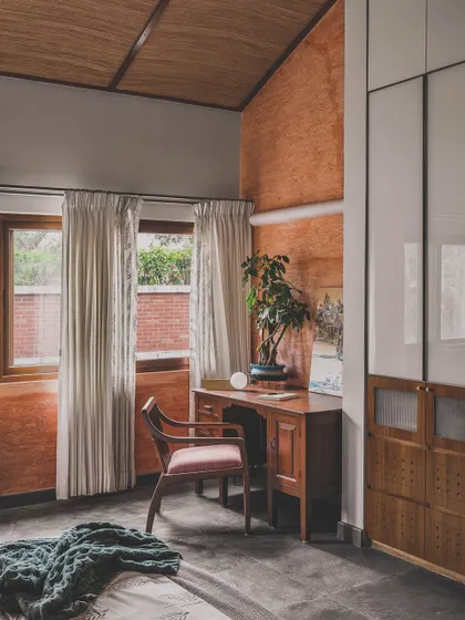 This bedroom study corner combines rustic and modern elements, with an exposed terracotta wall, a vaulted ceiling, and sleek white wardrobes. It’s a warm, personal space designed for work and rest.
