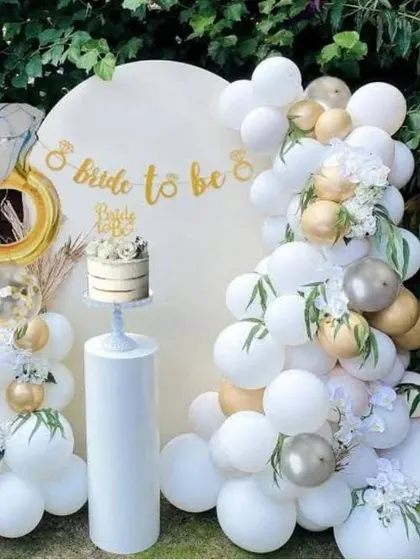 An elegant outdoor bridal shower setup in white and gold. This decor features a white circular backdrop, a white balloon garland with gold accents, a 'Bride to be' banner, and a large diamond ring foil balloon.