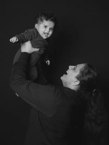 A joyful black and white photo of a mother lifting her laughing baby high in the air against a dark background.