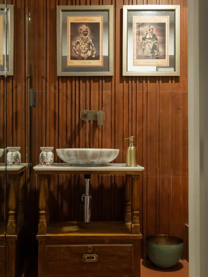 This bathroom exudes a rich warmth with wooden paneling, an antique counter, and a marble basin. The bronze tap complements the frames of the old Indian prints, creating a space with timeless, heritage appeal.