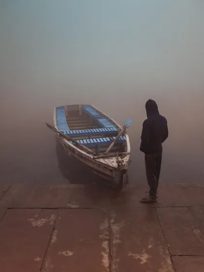 A person stands by a boat on a foggy morning at the Yamuna Ghat. The mood is quiet, contemplative, and solitary.