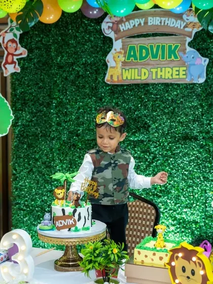 The birthday boy, dressed in a camouflage vest, stands by his jungle-themed cake table. This candid shot captures a moment of exploration during his 'Wild Three' birthday party.