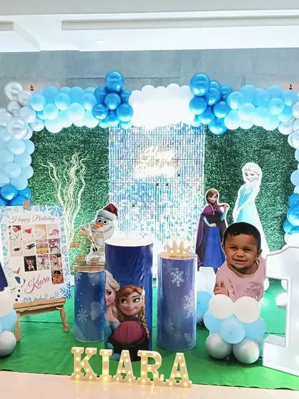 A Frozen-themed first birthday party with a grass-like backdrop and a silver sequin panel. The setup includes character cutouts and a milestone photo board.