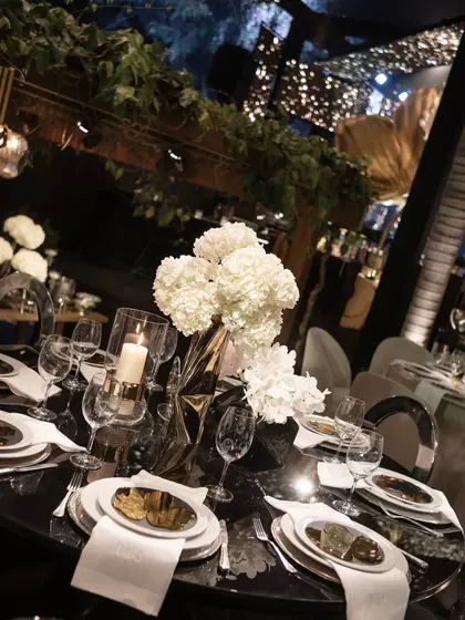 A glamorous table setting with reflective surfaces that catch the light beautifully. The centerpiece of white hydrangeas in a geometric gold vase adds a modern, sculptural element.