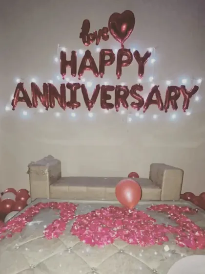 A simple yet heartfelt anniversary decoration. The wall features "Happy Anniversary" foil balloons with lights, and the bed is decorated with rose petals.