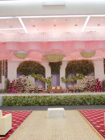 A wide view of a pastel-themed reception stage. The design incorporates soft pink draping and large floral installations, creating a dreamy and romantic setting for the couple.