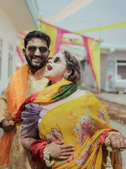 A candid shot of a couple laughing and having fun outdoors during their haldi or mehendi function, showcasing their vibrant outfits and joyful interaction.
