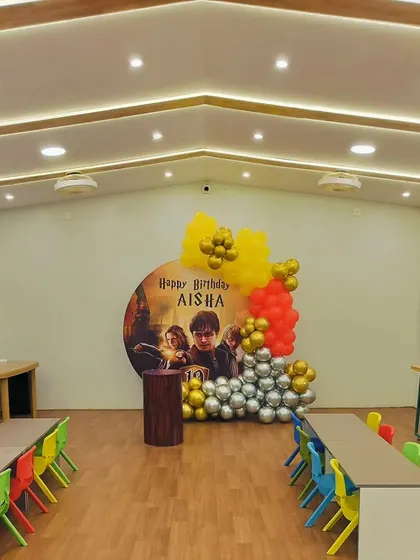 A Harry Potter themed birthday party set up in a large hall. The decor features a custom circular backdrop with characters from the movie, surrounded by balloons in house colors.