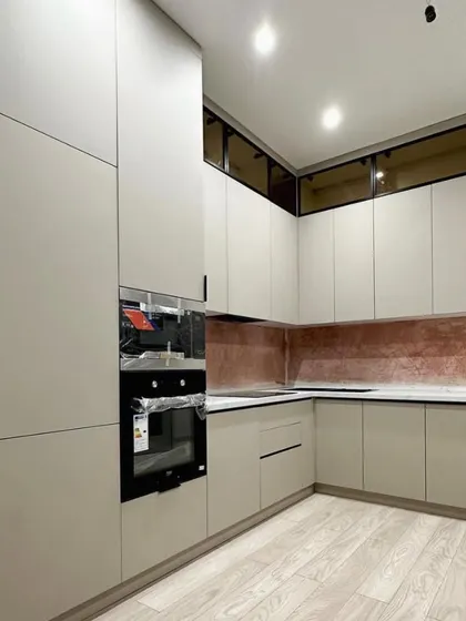 This elegant kitchen in Palam Vihar features matte beige handle-less cabinets and a striking rose-gold textured backsplash. The upper cabinets with tinted glass add a layer of sophistication.
