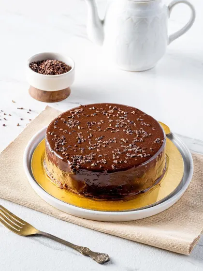 A whole double chocolate cake with a glossy ganache finish, sprinkled with chocolate shavings. The simple, elegant presentation on a white marble surface makes the cake the undeniable star of the photo.