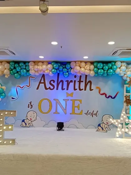 An "Ashrith is ONEderful" theme for a first birthday party. The stage is decorated with a large backdrop, marquee letters spelling "ONE," and cute baby illustrations.