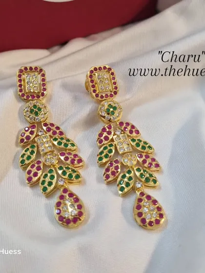 A clear, detailed studio shot of the "Charu" gold-plated Jadau earrings. The design features red and green stones, making them a festive and traditional choice for weddings.