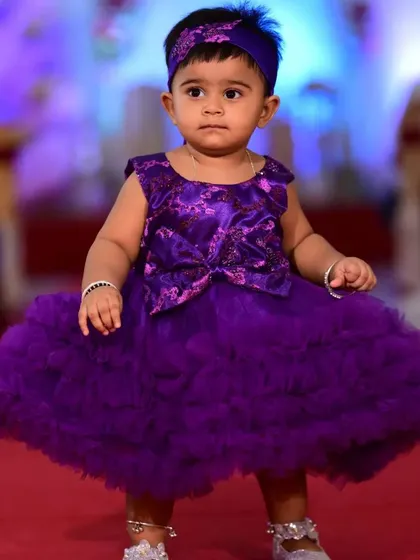 This little one is ready to celebrate in her vibrant purple party dress. The design features a sequined and patterned bodice with a large bow detail, flowing into a very full, multi-layered ruffled tulle skirt.