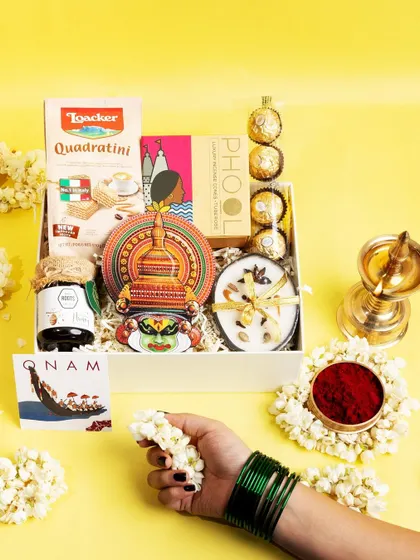 A hand adorned with green bangles reaches towards the 'Jamanthi Poo' hamper, adding a human touch and evoking the festive spirit of Onam.