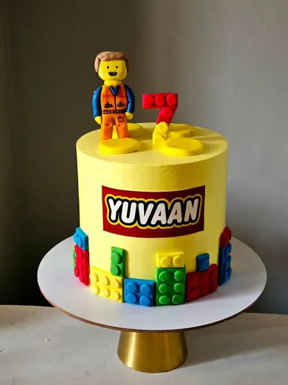 This bright and cheerful Lego-themed cake is perfect for a young builder. It features a handcrafted Lego man figurine, edible Lego blocks around the base, and the iconic Lego logo with a personalized name.