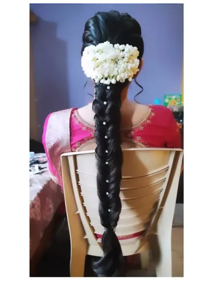 A neat fishtail braid with pearl accents, finished with a fresh jasmine gajra, seen from a seated perspective.