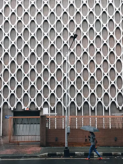 A person with an umbrella walks past a building with a striking geometric facade. The contrast between the organic human figure and the rigid, repeating pattern of the wall makes for a compelling minimalist shot.