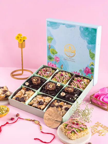 A Raksha Bandhan hamper box filled with assorted brownies and sweets, styled against a soft pink background with a rakhi and flowers.