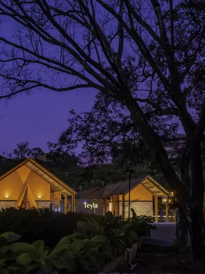 Teyla at dusk, with warm light spilling from its gabled structures. The building's silhouette against the evening sky creates a welcoming beacon for visitors, while the surrounding landscape is subtly lit.