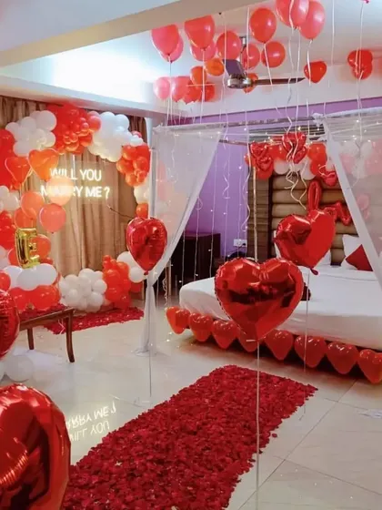 A "Will You Marry Me?" proposal setup. This romantic room decoration features a red rose petal aisle, a balloon arch, a neon sign, and a bed decorated with heart-shaped balloons.