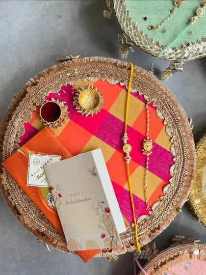 A stunning handmade Rakhi thal featuring a vibrant pink and orange checkered fabric, gota patti border, and a matching Rakhi set. It comes complete with a diya and containers for Roli and Chawal.