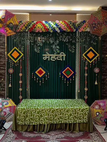 An elegant Mehendi stage with a dark green backdrop, colorful umbrellas, and traditional hangings, perfect for indoor banquet hall events.