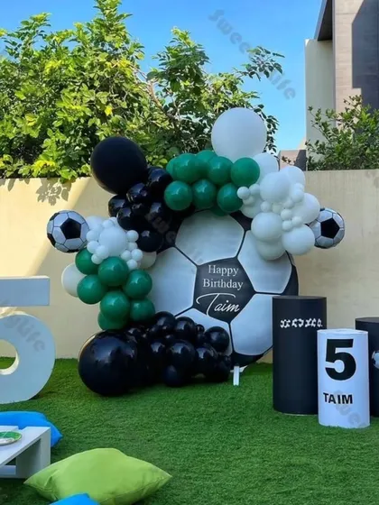 A cool football-themed 5th birthday party. This outdoor setup includes a giant football backdrop, a green, black, and white balloon garland, and a large marquee number '5'.