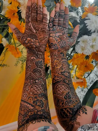 A beautiful shot of a full bridal henna against a floral backdrop. The design features bold leaf motifs and intricate geometric patterns, covering the arms completely for a stunning effect.