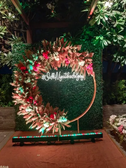 A beautiful photo booth with a custom neon hashtag. The circular frame is decorated with pampas grass and red flowers against a green wall, making it a stylish spot for pictures.