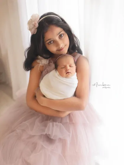 A beautiful, light-filled portrait of an older sister proudly holding her swaddled newborn sibling. Her gentle smile speaks volumes about her love for the new baby.
