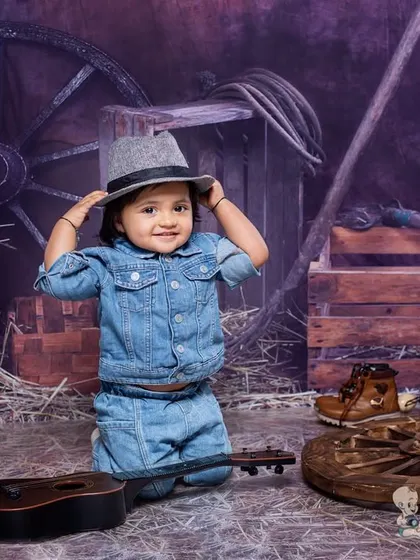 A toddler in a denim outfit and fedora hat, kneeling in a rustic barn setting. The hay, wagon wheels, and guitar prop create a charming country-western theme.